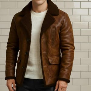 May include: A brown leather jacket with a shearling collar and trim. The jacket features a button-up front, two front pockets, and a classic design. The jacket is worn over a white sweater.