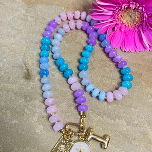 Puede incluir: Un collar de cuentas con un degradado de colores, incluyendo cuentas rosas, moradas y azules. El collar tiene colgantes dorados, incluyendo un colgante rectangular con un diseño de sol, un colgante redondo con una estrella de mar y un colgante con forma de la letra "I". Una margarita rosa está en el fondo.