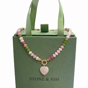 Puede incluir: Un collar con un colgante de corazón rosa y cuentas multicolores se muestra en una caja de regalo verde. La caja tiene el texto "STONE & ASH" impreso. Las cuentas del collar son rosas, verdes y rojas.