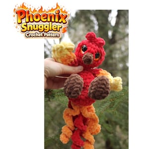 May include: A handmade crochet phoenix toy in red, yellow, and orange. The plush bird has brown feet and a tail, with the text "Phoenix Snuggler Crochet Pattern" in a flame-like design. The toy is held against a blurred green background.