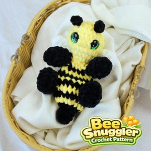 Puede incluir: Un juguete de abeja hecho a mano con rayas amarillas y negras, extremidades negras y difusas y ojos verdes. La abeja está en una cesta de mimbre sobre una tela blanca. La imagen incluye el texto "Bee Snuggler Crochet Pattern".