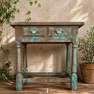 Puede incluir: Una mesa auxiliar rectangular de madera con dos cajones, con un acabado desgastado en tonos de cobre oxidado, verde azulado y bronce. La mesa tiene cuatro patas torneadas conectadas por un travesaño inferior y dos tiradores de metal.