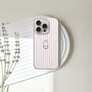 May include: A phone case with a clear border and a pink and white striped design. The case features a cartoon bunny illustration in the center. The phone case is resting on a white plate.