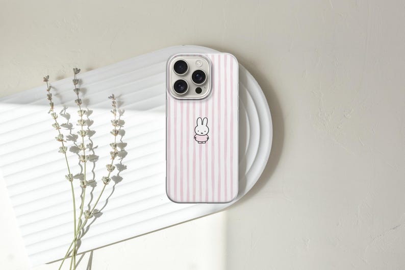 May include: A phone case with a clear border and a pink and white striped design. The case features a cartoon bunny illustration in the center. The phone case is resting on a white plate.
