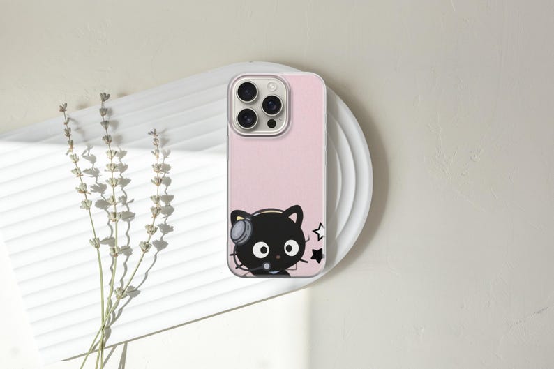 Op de afbeelding: Een roze telefoonhoesje met een zwarte kat met een koptelefoon en sterren. Het telefoonhoesje ligt op een wit bord met gedroogde bloemen links.