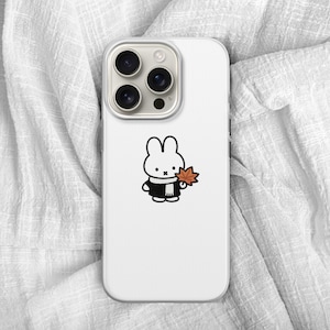 May include: White phone case featuring a cartoon rabbit character wearing a black coat and scarf, holding an orange leaf. The phone case has a triple-lens camera and is set against a white, textured background.