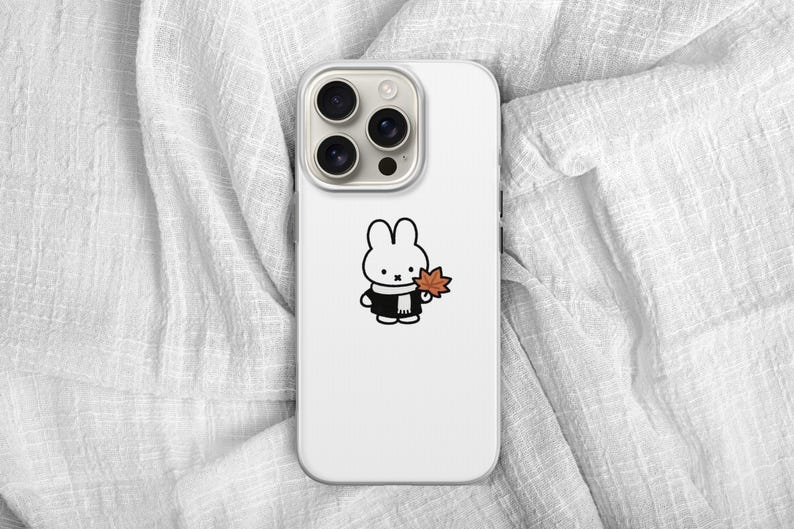May include: White phone case featuring a cartoon rabbit character wearing a black coat and scarf, holding an orange leaf. The phone case has a triple-lens camera and is set against a white, textured background.