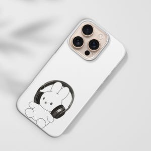 May include: White phone case featuring a black and white illustration of a cartoon bunny wearing headphones. The phone case has a clear camera lens protector and is angled on a white surface.