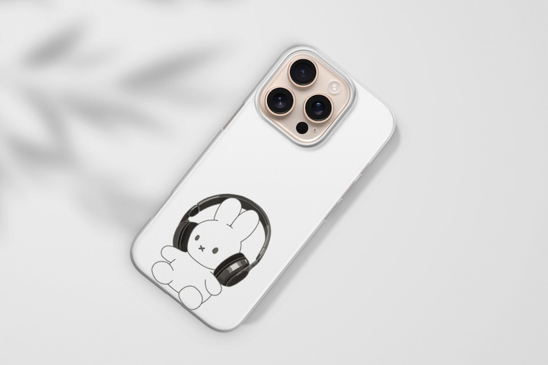 May include: White phone case featuring a black and white illustration of a cartoon bunny wearing headphones. The phone case has a clear camera lens protector and is angled on a white surface.