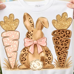 May include: White sweatshirt featuring an Easter-themed design. The design includes a brown bunny with a pink bow, two carrots with gold glitter tops, and a floral accent. The bunny and carrots have a leopard print pattern.