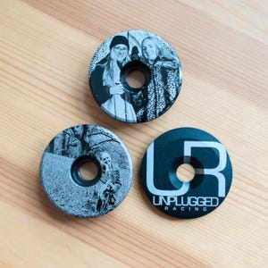 May include: Three circular bicycle headset caps with a centre hole. One cap features a black and white photograph of two people, another shows a black and white outdoor portrait of a person, and the third is black with the white text UNPLUGGED RACING.