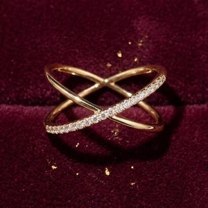 Puede incluir: Un anillo dorado con un dise&ntilde;o cruzado, adornado con una hilera de peque&ntilde;as piedras brillantes. El anillo se encuentra sobre un fondo burdeos oscuro, con algunas motas doradas. El dise&ntilde;o del anillo presenta dos bandas que se cruzan.
