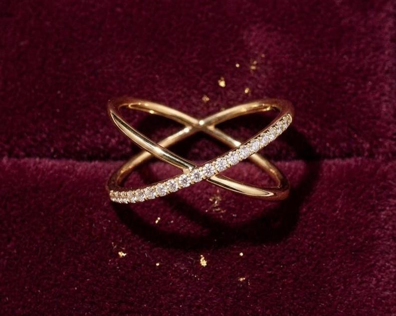 Puede incluir: Un anillo dorado con un dise&ntilde;o cruzado, adornado con una hilera de peque&ntilde;as piedras brillantes. El anillo se encuentra sobre un fondo burdeos oscuro, con algunas motas doradas. El dise&ntilde;o del anillo presenta dos bandas que se cruzan.