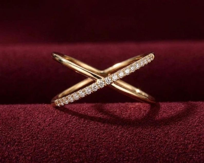 Puede incluir: Un anillo dorado con dise&ntilde;o cruzado y una fila de peque&ntilde;as gemas claras engastadas en una de las bandas. El anillo se exhibe sobre una superficie de terciopelo rojo oscuro, creando una sensaci&oacute;n lujosa. El dise&ntilde;o del anillo es moderno y elegante.