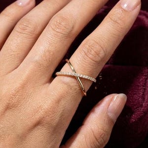 Puede incluir: Un anillo dorado entrecruzado con peque&ntilde;as piedras preciosas transparentes engarzadas a lo largo de una banda. El anillo se lleva en un dedo, con la mano y parte del fondo visibles.