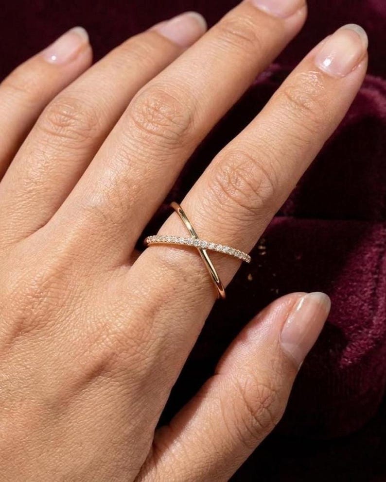 Puede incluir: Un anillo dorado entrecruzado con peque&ntilde;as piedras preciosas transparentes engarzadas a lo largo de una banda. El anillo se lleva en un dedo, con la mano y parte del fondo visibles.