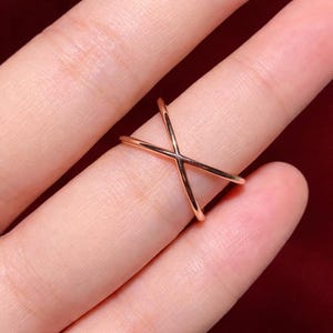 Puede incluir: Un anillo de oro rosa con un dise&ntilde;o en forma de X. El anillo se lleva en un dedo, mostrando su estilo minimalista y moderno. La superficie pulida del anillo refleja la luz, resaltando su elegante simplicidad.