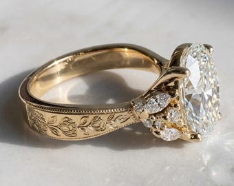 Könnte beinhalten: Ein Goldring mit einem großen oval geschliffenen Mittelstein, akzentuiert durch kleinere marquise-geschliffene Steine an der Seite. Die Ringschiene ist aus Gelbgold gefertigt und zeigt eine florale Gravur mit Milgrain-Details.