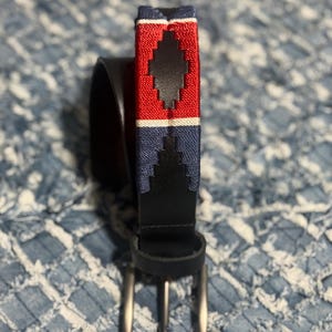 Op de afbeelding: Een zwarte leren riem met een geweven textielgedeelte met een herhalend ruitpatroon in rood, wit en marineblauw. De riem is voorzien van een zilverkleurige metalen gesp en een bijpassende leren lus, wat zorgt voor een klassiek ontwerp.
