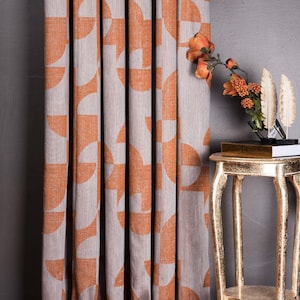 Könnte beinhalten: Vorhänge mit einem geometrischen Muster in Orange und Grau. Die Vorhänge hängen vor einer dunkelgrauen Wand. Rechts neben den Vorhängen steht ein kleiner goldener Beistelltisch mit Blumenarrangement.