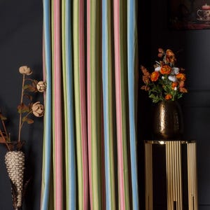 May include: Curtain panel with vertical stripes in pastel shades of blue, pink, and green. The curtain hangs against a dark wall. A gold-coloured side table with a vase of flowers is to the right.