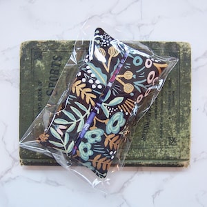 Może przedstawiać: Pi&oacute;rnik w kwiatowy wz&oacute;r w przezroczystym plastikowym etui. Etui ma ciemne tło ze złotymi, turkusowymi i r&oacute;żowymi wzorami kwiatowymi. Spoczywa na zabytkowej zielonej książce. Widoczny jest napis "SPORTS".