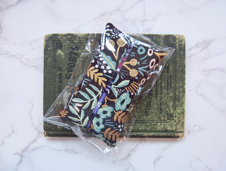 Może przedstawiać: Pi&oacute;rnik w kwiatowy wz&oacute;r w przezroczystym plastikowym etui. Etui ma ciemne tło ze złotymi, turkusowymi i r&oacute;żowymi wzorami kwiatowymi. Spoczywa na zabytkowej zielonej książce. Widoczny jest napis "SPORTS".