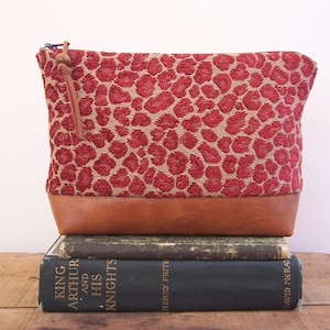 May include: A zippered pouch featuring a textured red and beige animal print pattern on the upper portion and a solid brown leather-like base. A thin brown leather cord with a small wooden bead is attached to the metal zipper pull at the top.