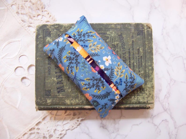 Może przedstawiać: Niebieska poszewka na pudełko chusteczek w kwiaty z otworem, spoczywająca na zielonej, zabytkowej książce. Poszewka ma wz&oacute;r kwiat&oacute;w i liści w odcieniach pomarańczu, bieli i zieleni. Na książce widnieje napis "MANIA".