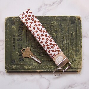 May include: A key wristlet with a brown leaf pattern on a white background, attached to a silver-colored metal clasp. The wristlet rests on a vintage green book with gold lettering, next to a brass key.