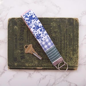 May include: A fabric key fob with a floral and plaid pattern in blue, white, and green. The key fob has a silver-colored metal ring and is next to a gold-colored key. The key fob is on top of a vintage green book.
