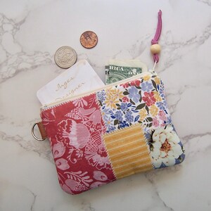 May include: A rectangular zippered pouch featuring a patchwork design of floral and striped fabric in shades of pink, yellow, blue, and white. The pouch includes a pink leather pull tab with a wooden bead and a metal D-ring on the side.