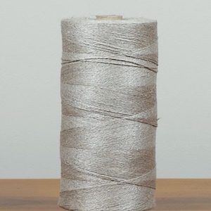 Peut inclure: Une bobine de fil gris clair, cylindrique et bien enroulée, est posée sur une surface en bois. Le fil semble être fait d'une fibre naturelle, avec de subtiles variations de couleur et de texture. Idéal pour les projets de couture et d'artisanat.