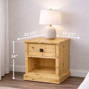 May include: A light-colored wooden nightstand with a drawer and open shelf. The nightstand is 24 inches tall, 21 3/4 inches wide, and 18 1/2 inches deep. A white lamp with a beige base sits on top. The drawer has a black square handle.