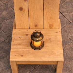 May include: A small, light-colored wooden chair with a backrest made of three vertical planks. A decorative lantern with a lit candle sits on the chair's seat. The lantern is bronze-colored with star-shaped cutouts.