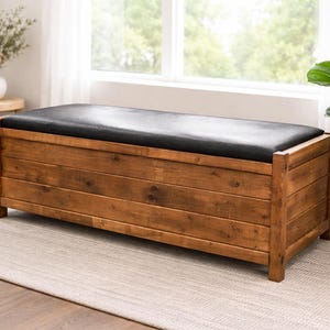 Puede incluir: Un banco de almacenamiento de madera con una parte superior acolchada negra. El banco está hecho de madera de color marrón claro con vetas visibles y tiene forma rectangular. Se encuentra sobre una alfombra de color claro frente a una ventana.