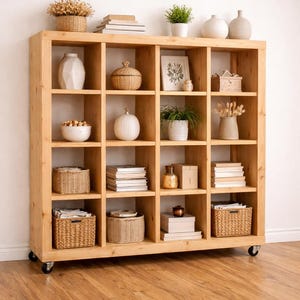 May include: A light-colored wooden bookcase with multiple cubby shelves. The shelves are filled with decorative items like plants, books, vases, and woven baskets. The bookcase sits on wheels and is against a white wall.