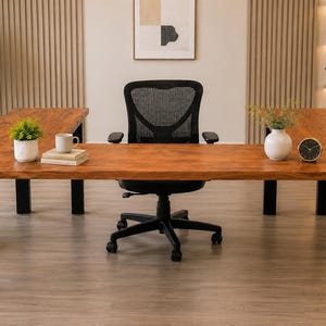 Puede incluir: Un escritorio de madera grande en forma de U con acabado marrón cálido y patas de metal negro. El escritorio incluye una silla de oficina de malla negra, una pequeña planta en maceta, una taza blanca sobre libros, un jarrón blanco y una lámpara.