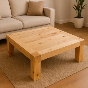 May include: A square, light-colored wooden coffee table with a natural finish. The table has a simple, modern design with thick legs and a sturdy top. It sits on a woven rug in a living room setting, with a beige sofa and a potted plant in the background.