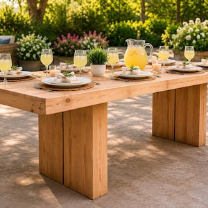 May include: A large, rectangular wooden dining table set for a meal outdoors. The table is made of light-colored wood and has a pitcher of yellow liquid, glasses, plates, and small potted plants. The table is set on a patio.