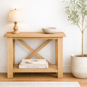 May include: A light oak wood console table with an X-shaped support. A beige lamp with a linen shade sits on the top, along with a speckled bowl. A stack of books and a small bowl are on the lower shelf. A potted plant is to the right.
