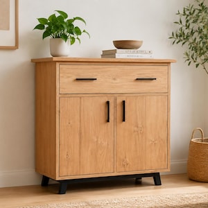 May include: A light brown wooden cabinet with a drawer and two doors. The cabinet has black handles and legs. A small potted plant, a wooden bowl, and books sit on top. A woven basket with a plant is next to the cabinet.