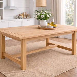 Puede incluir: Una mesa de comedor de madera clara con una superficie rectangular y patas robustas. La mesa está sobre una alfombra beige en una cocina luminosa. Un jarrón con flores blancas está sobre la mesa, de unos 150 cm de largo.