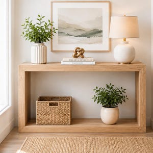 May include: A light wood console table with a framed landscape print, a lamp, and decorative items. The table holds a white vase with greenery, a woven basket, and a potted plant. A neutral rug is on the floor.