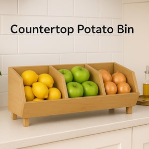 Op de afbeelding: Een houten aardappelbak op het aanrecht gevuld met citroenen, groene appels en uien. De bak heeft drie compartimenten en staat op een wit aanrecht. Links van de bak staat een kleine plant in een pot.