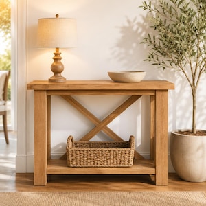 Pode incluir: Uma mesa consola de carvalho claro com uma estrutura de suporte em forma de X, com um tampo retangular e uma prateleira inferior. Sobre a mesa, uma luminária de madeira com cúpula bege, uma tigela de pedra rasa e um cesto de vime retangular.