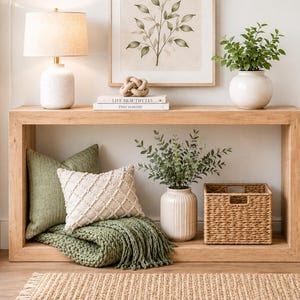 Könnte beinhalten: Ein Konsolentisch aus hellem Holz mit einer Lampe, Kunstwerken und dekorativen Gegenständen. Der Tisch trägt eine weiße Lampe, einen gerahmten botanischen Druck und eine Vase mit Grün. Darunter befinden sich Kissen, eine Decke, eine Vase und ein Weidenkorb. Auf dem Boden liegt ein Jute-Teppich.
