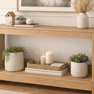 Puede incluir: Una consola de madera clara con una impresión de paisaje enmarcada encima. La mesa está decorada con una pequeña figura de casa, plantas en macetas, velas, libros y un jarrón con flores secas. La estética general es minimalista y natural.
