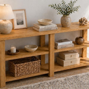 May include: A light-colored wooden console table with three shelves. The top shelf holds a lamp, framed art, books, and decorative bowls. The lower shelves display a woven basket, candles, and decorative boxes. A vase with greenery and a wooden knot sit on the top shelf.