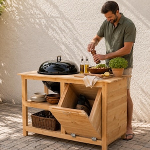 Könnte beinhalten: Eine hölzerne Outdoor-Grillstation mit einem schwarzen Grill, Stauraum und einer Arbeitsfläche. Die Station hat ein unteres Regal mit einem Korb und Schalen. Eine Person würzt einen Salat. Die Station ist aus hellem Holz gefertigt.